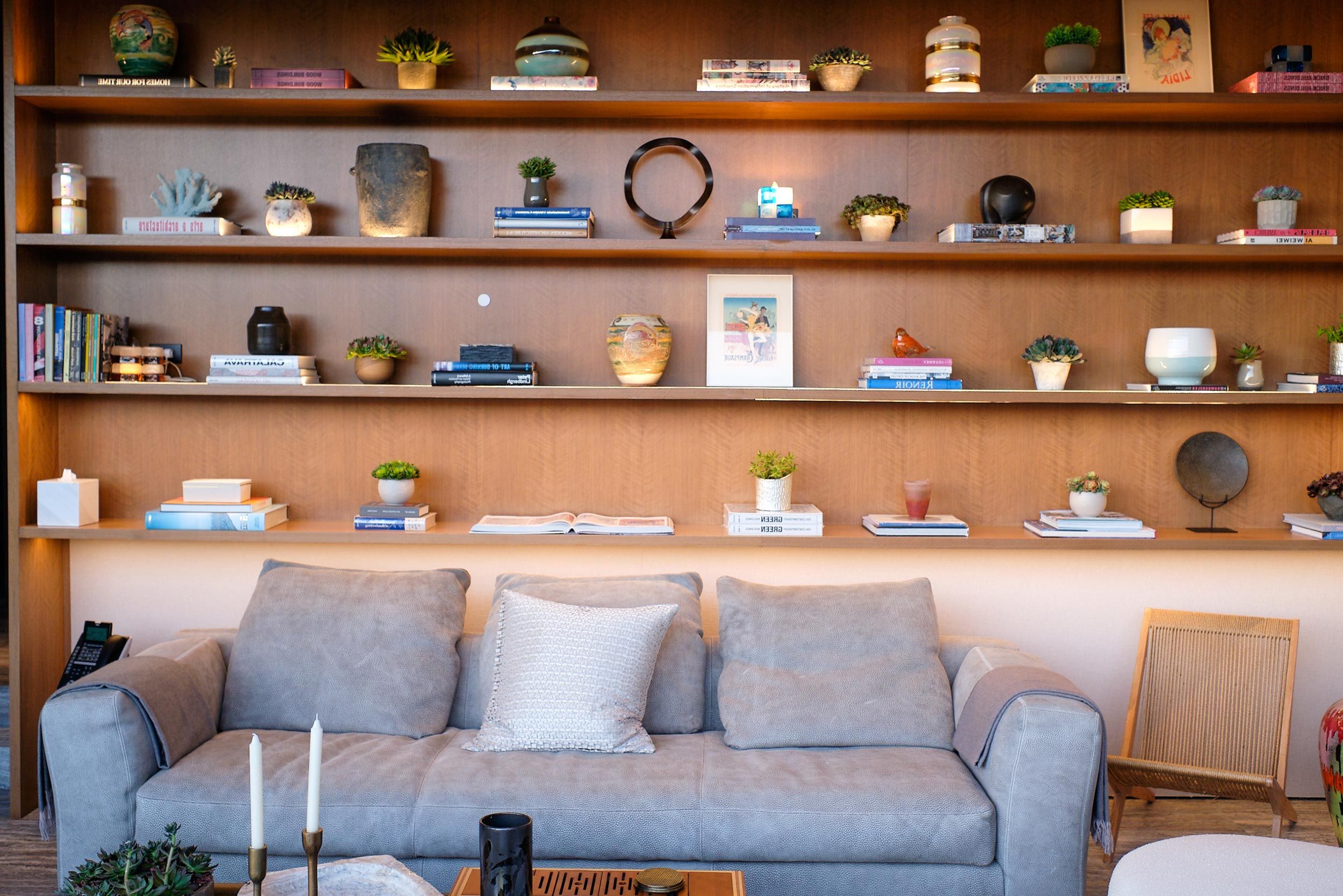 Cozy living room with a gray sofa, decorative pillows, and a wooden bookshelf filled with books, plants, and various decor items.