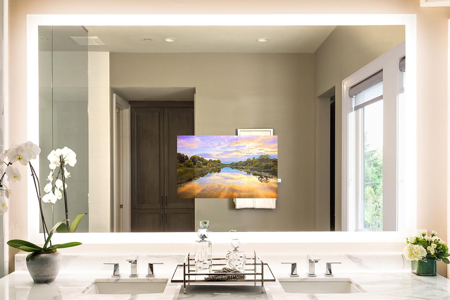 Elegant bathroom with a backlit mirror and a hidden TV screen, showcasing a serene landscape image, with a sleek double sink vanity and decorative orchids.